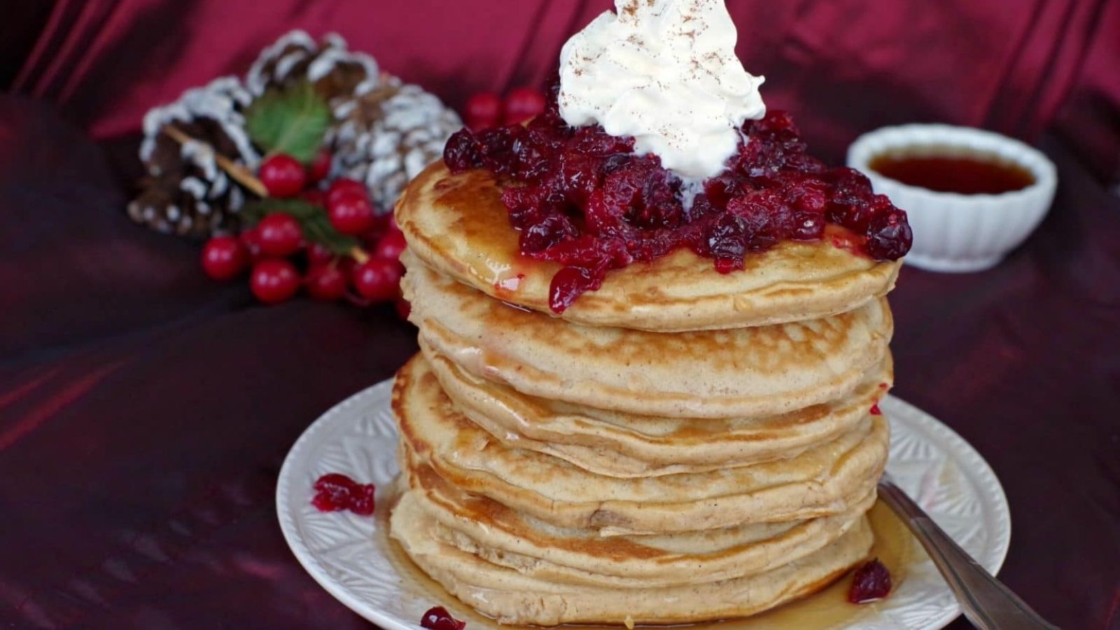 A stack of pancakes topped with cranberry sauce and whipped cream on a white plate, with a cup of syrup and festive decorations in the background.