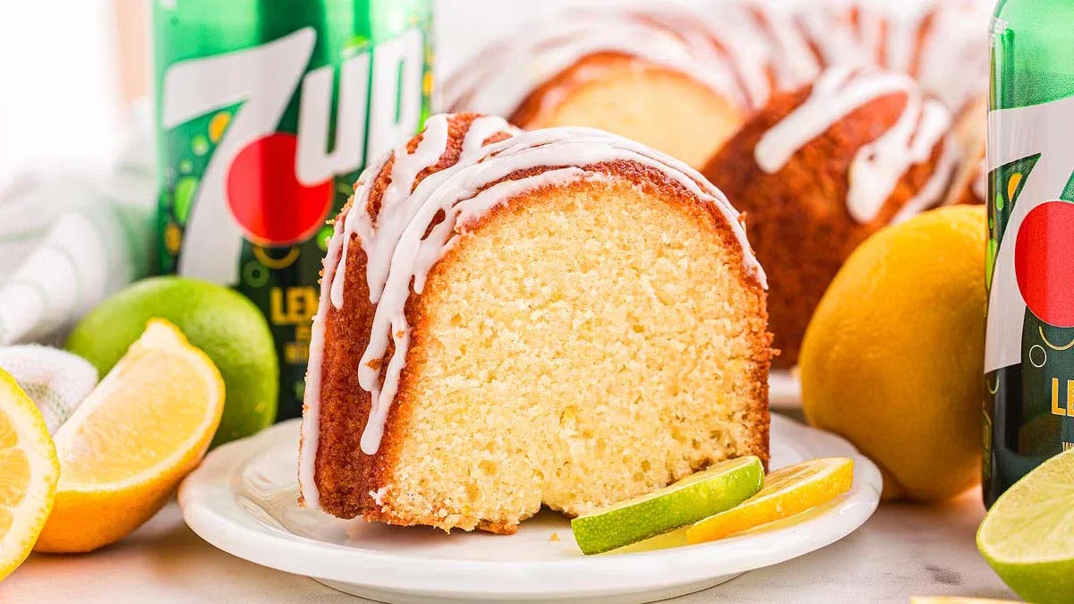 A slice of Bundt cake with white icing on a plate, garnished with lime and lemon slices, with 7UP cans and more cake in the background.