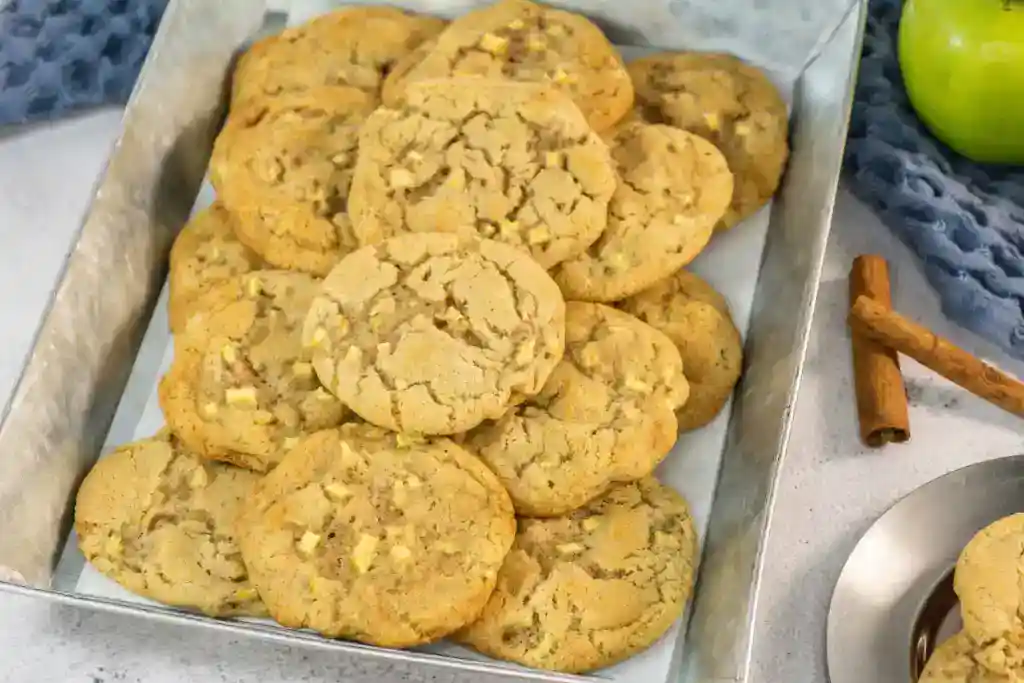 A metal tray filled with a pile of apple chunk cookies sits on a white surface next to cinnamon sticks and a green apple.