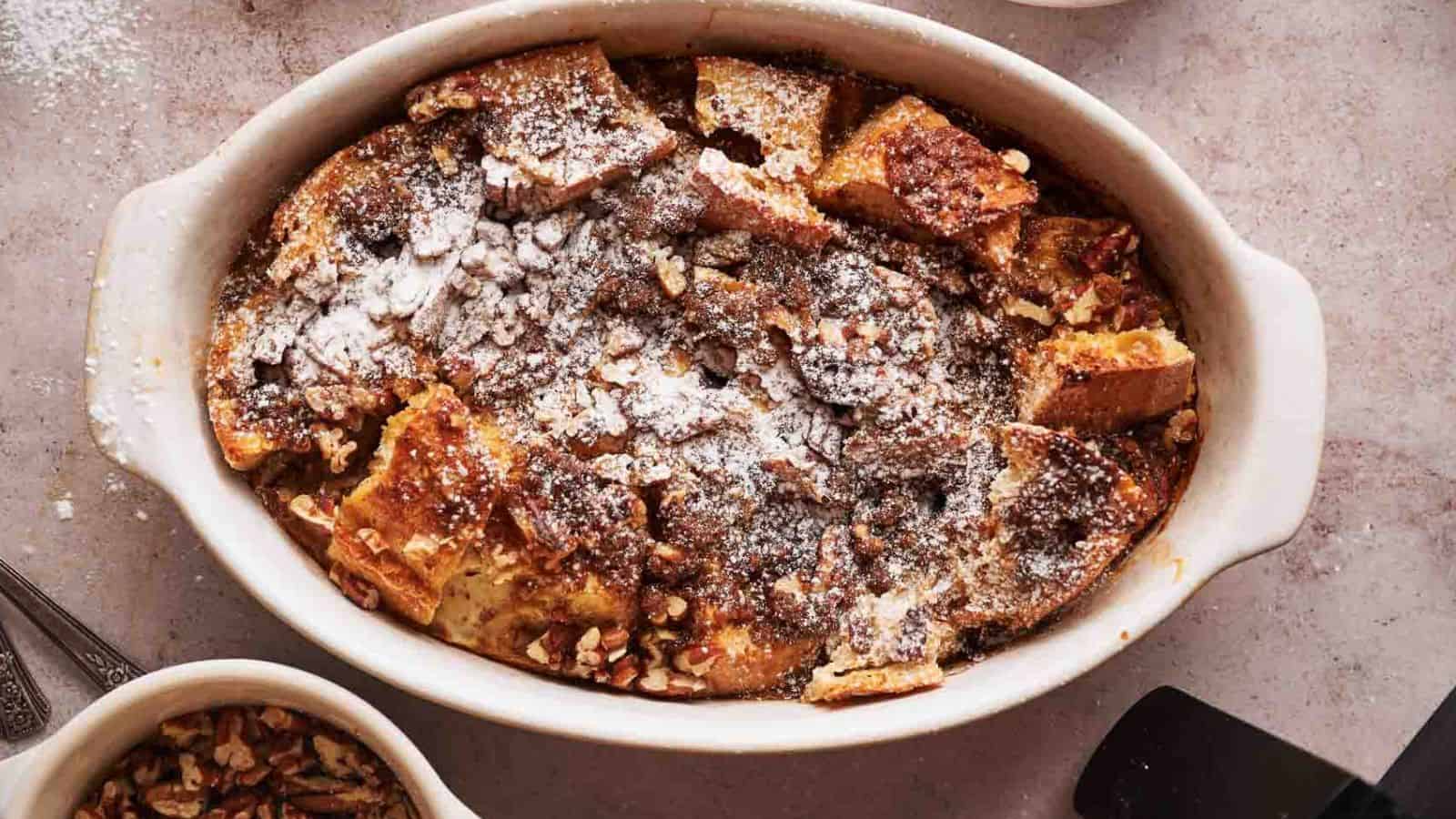 A baked bread pudding in a white oval dish, topped with powdered sugar and chopped nuts, sits on a light-colored surface.