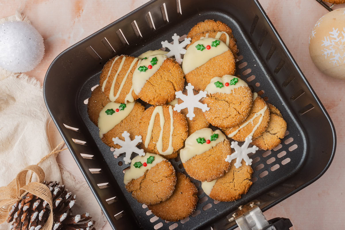 A batch of round cookies, some dipped in white chocolate and decorated with snowflake shapes and small green and red sprinkles, arranged in an air fryer basket.