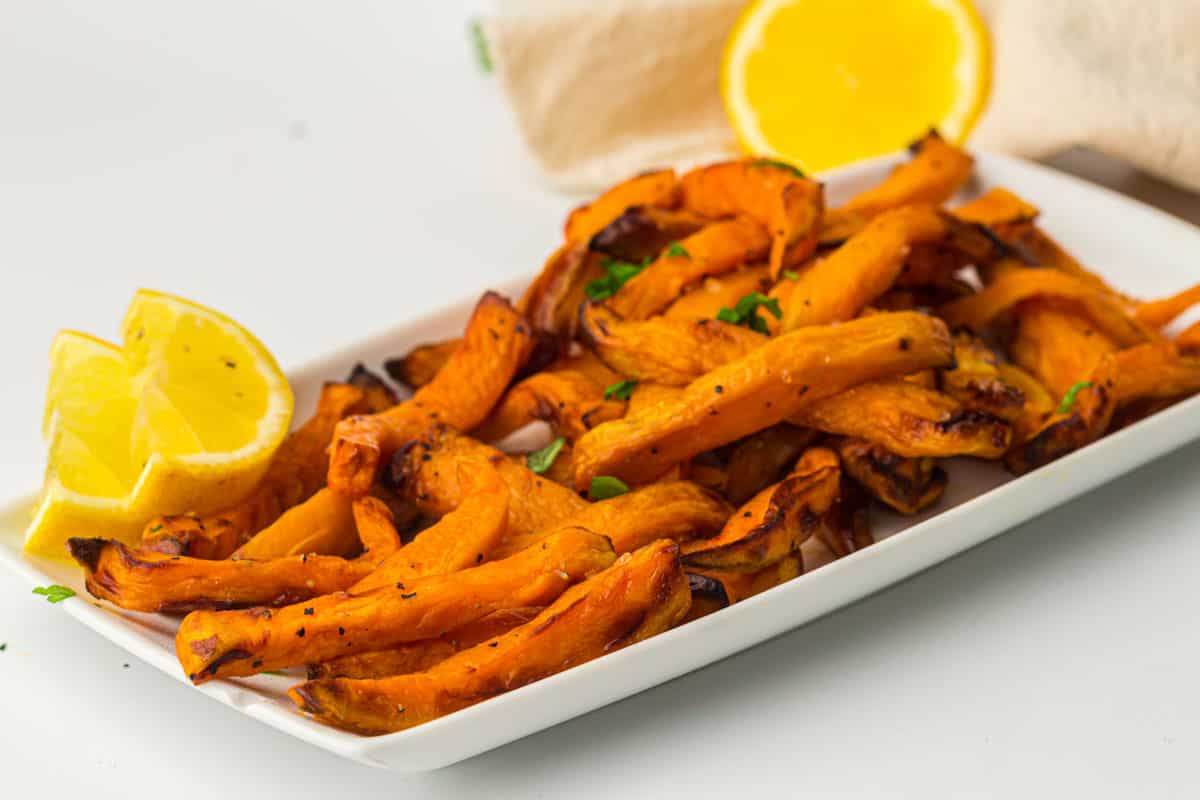 A rectangular white plate with roasted sweet potato fries garnished with parsley, served alongside lemon wedges.