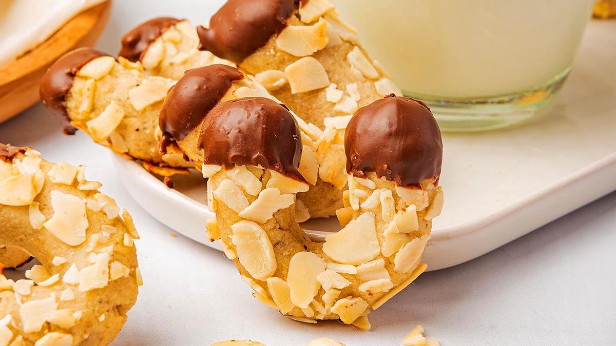 Almond crescent cookies partially dipped in chocolate are arranged on a white plate next to a glass of milk.