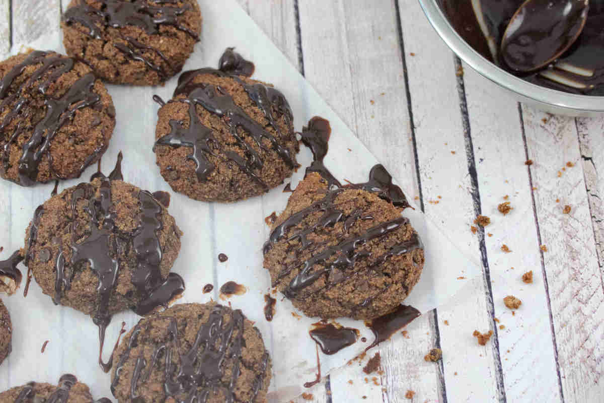 Chocolate cookies with a drizzle of chocolate icing are arranged on parchment paper, with crumbs scattered nearby and a bowl of chocolate sauce visible in the corner.