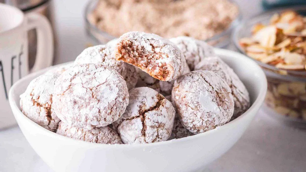 A white bowl filled with round, powdered sugar-coated cookies, with one cookie showing a bite taken out, placed near bowls of brown sugar and sliced almonds.