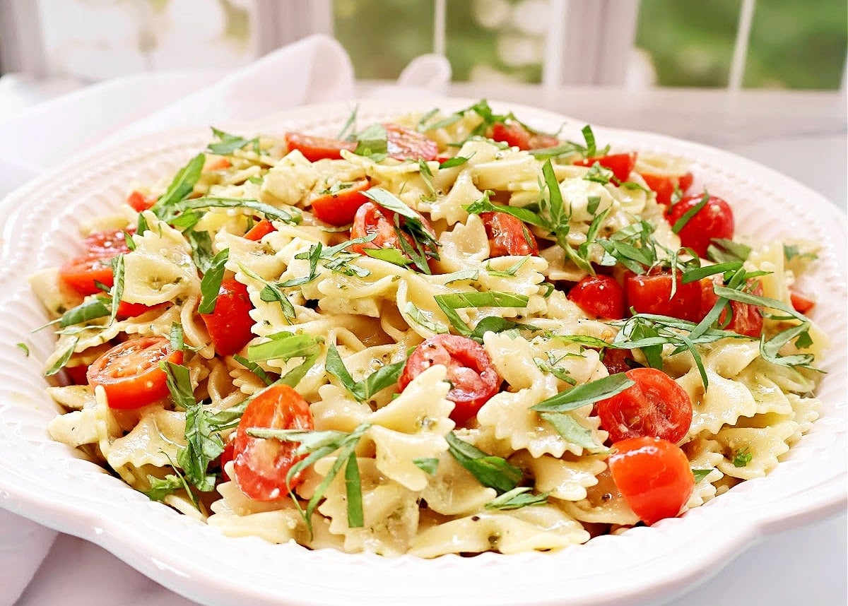 A white plate filled with bow-tie pasta, halved cherry tomatoes, green herbs, and a light dressing, set on a table by a window.