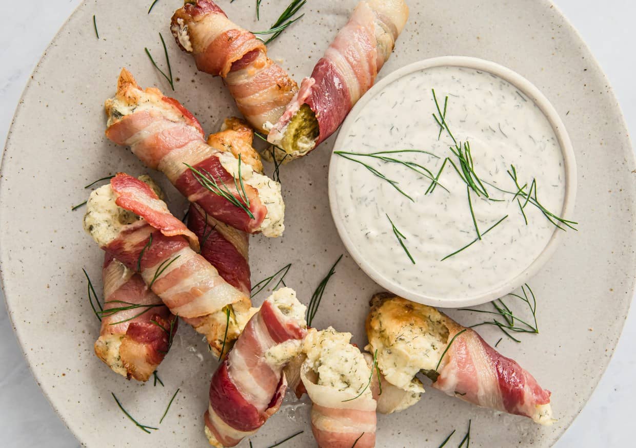 Bacon-wrapped stuffed chicken tenders are arranged on a plate with fresh dill and served with a bowl of creamy ranch dipping sauce.