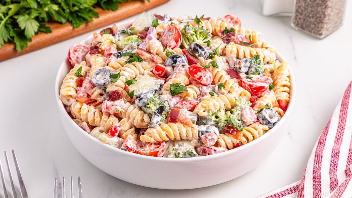 A bowl of rotini pasta salad with cherry tomatoes, black olives, broccoli, red onion, and a creamy dressing, garnished with parsley.