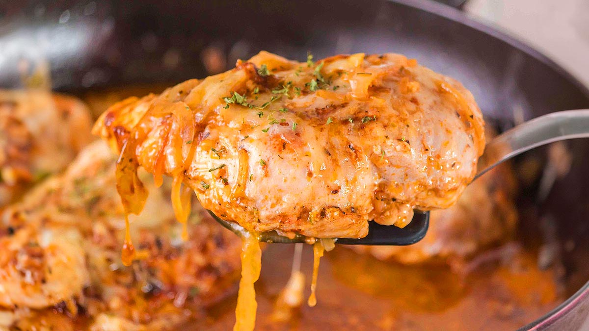 A cooked chicken breast covered in melted cheese and sauce is lifted from a skillet with a spatula.