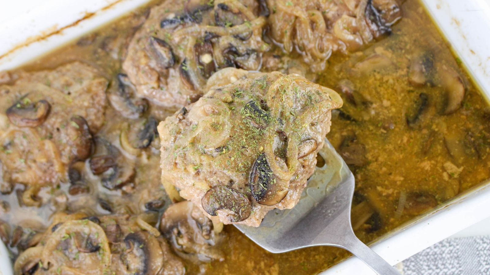 A serving spatula holds a portion of steak topped with mushrooms and onions above a baking dish containing several pieces in brown gravy.