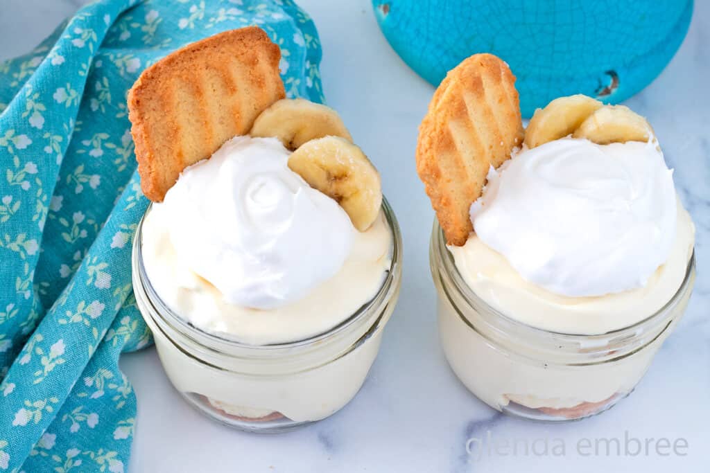 Two glass jars filled with banana pudding, topped with whipped cream, banana slices, and a cookie, placed on a white surface next to a blue patterned cloth.