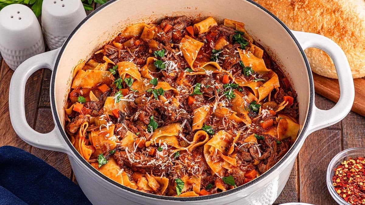 A white pot filled with pappardelle pasta, shredded beef, tomato sauce, vegetables, and grated cheese sits on a wooden table next to bread, salt and pepper shakers, and spice flakes.