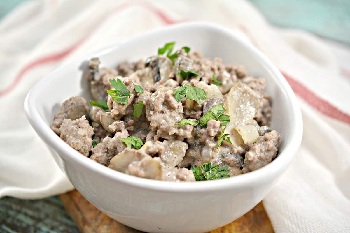 A white bowl filled with ground beef mixed with onions, mushrooms, and garnished with chopped parsley, set on a cloth napkin.