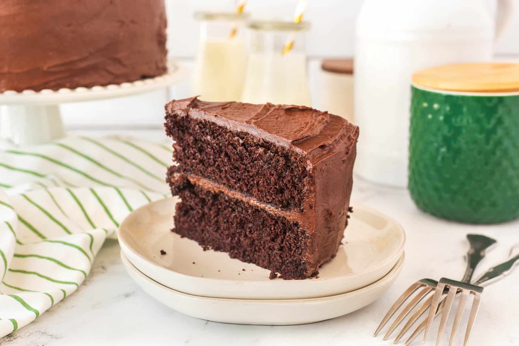 A slice of two-layer chocolate cake with chocolate frosting on a plate, with the rest of the cake, drinks, and utensils in the background.
