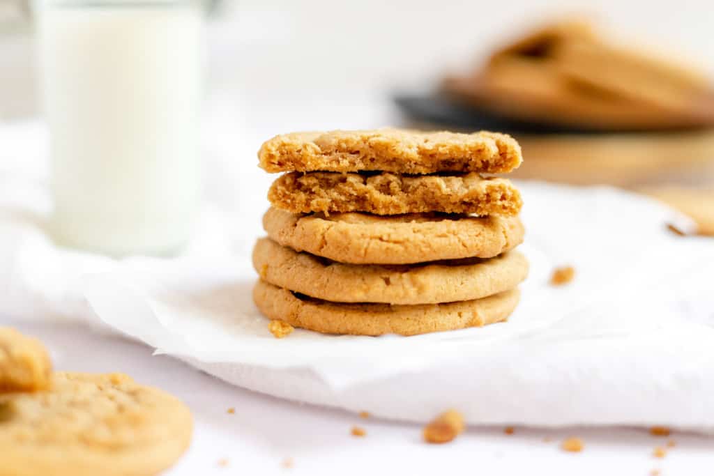 A stack of four peanut butter cookies on a white napkin, with the top cookie broken in half and a glass of milk in the background.