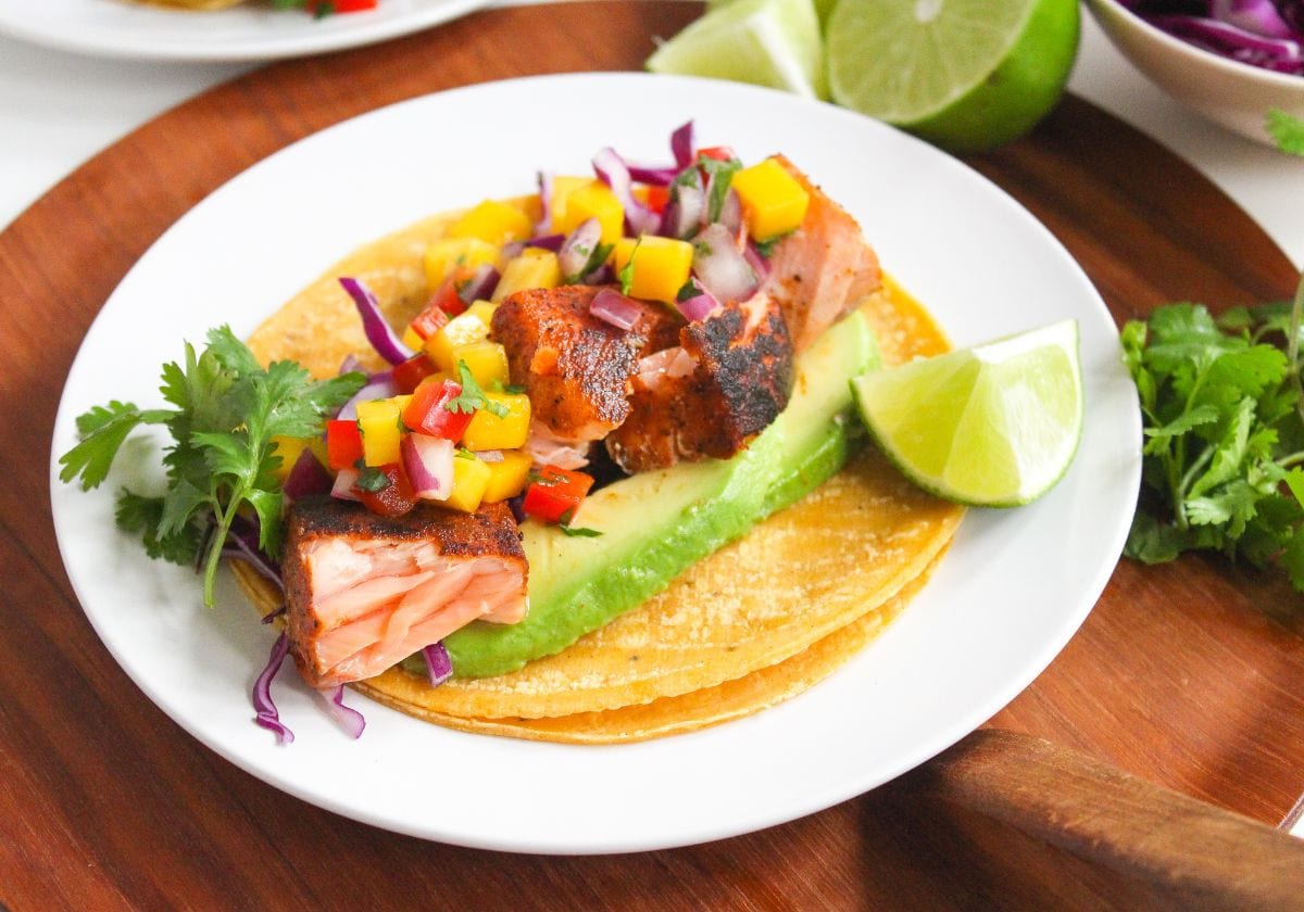 A plate with a salmon taco featuring avocado slices, red cabbage, mango salsa, cilantro, and a lime wedge on a wooden surface.
