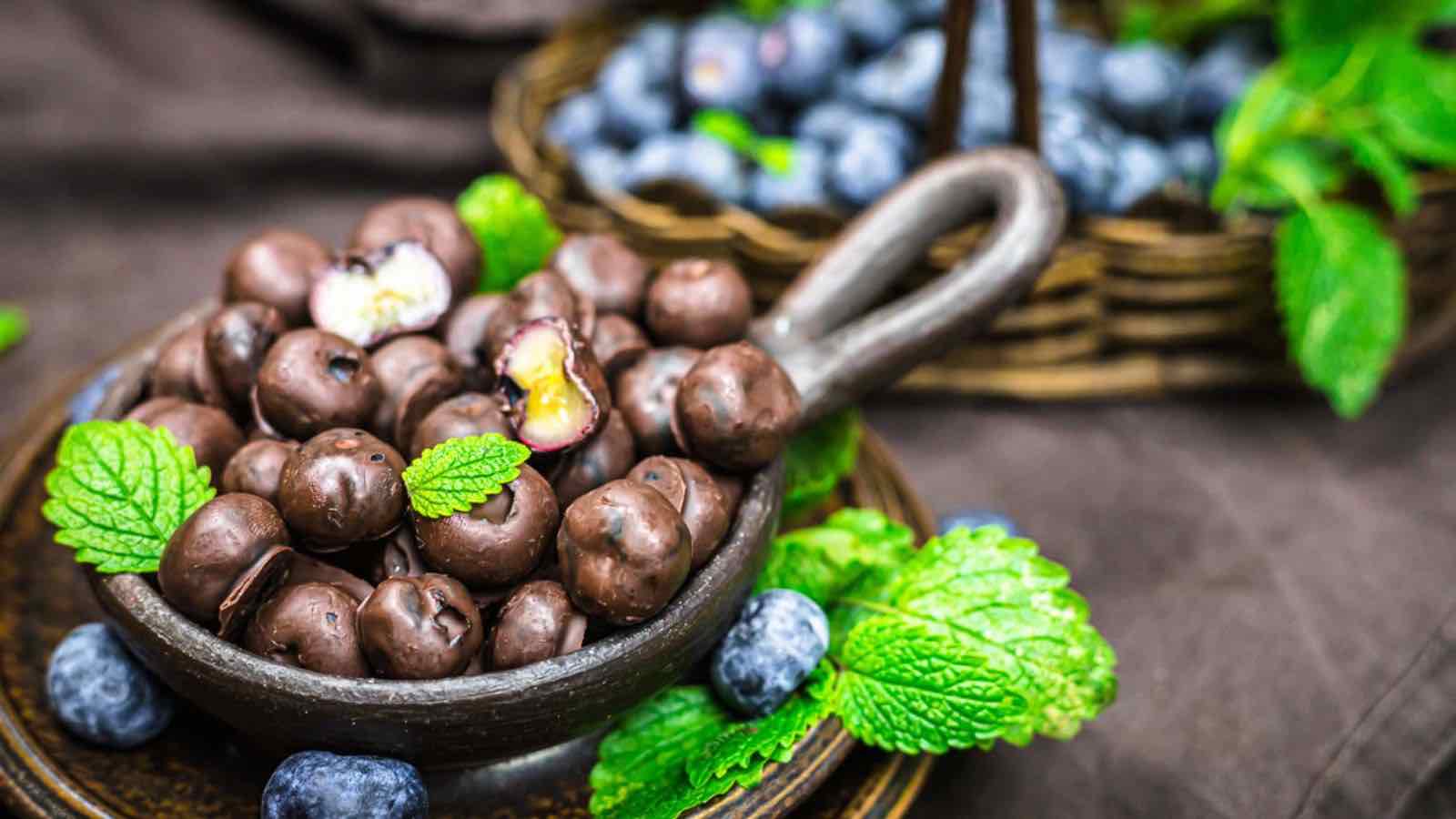 A bowl of chocolate-covered blueberries garnished with fresh mint leaves, with more blueberries and mint in the background.