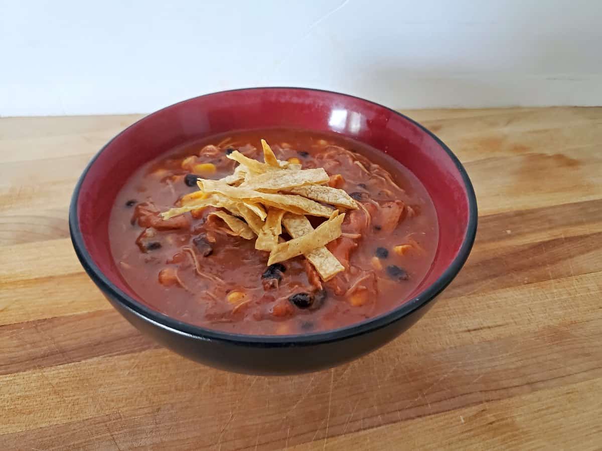 A bowl of tomato-based soup with black beans, corn, shredded chicken, and tortilla strips on top, placed on a wooden surface.