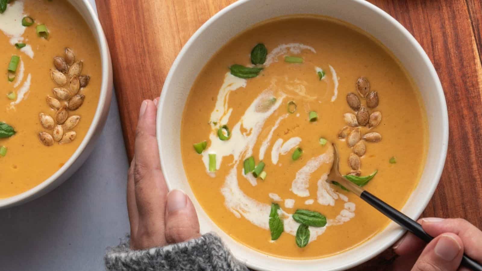 A hand holds a bowl of creamy orange soup garnished with pumpkin seeds, green onions, fresh herbs, and a swirl of cream, on a wooden surface.