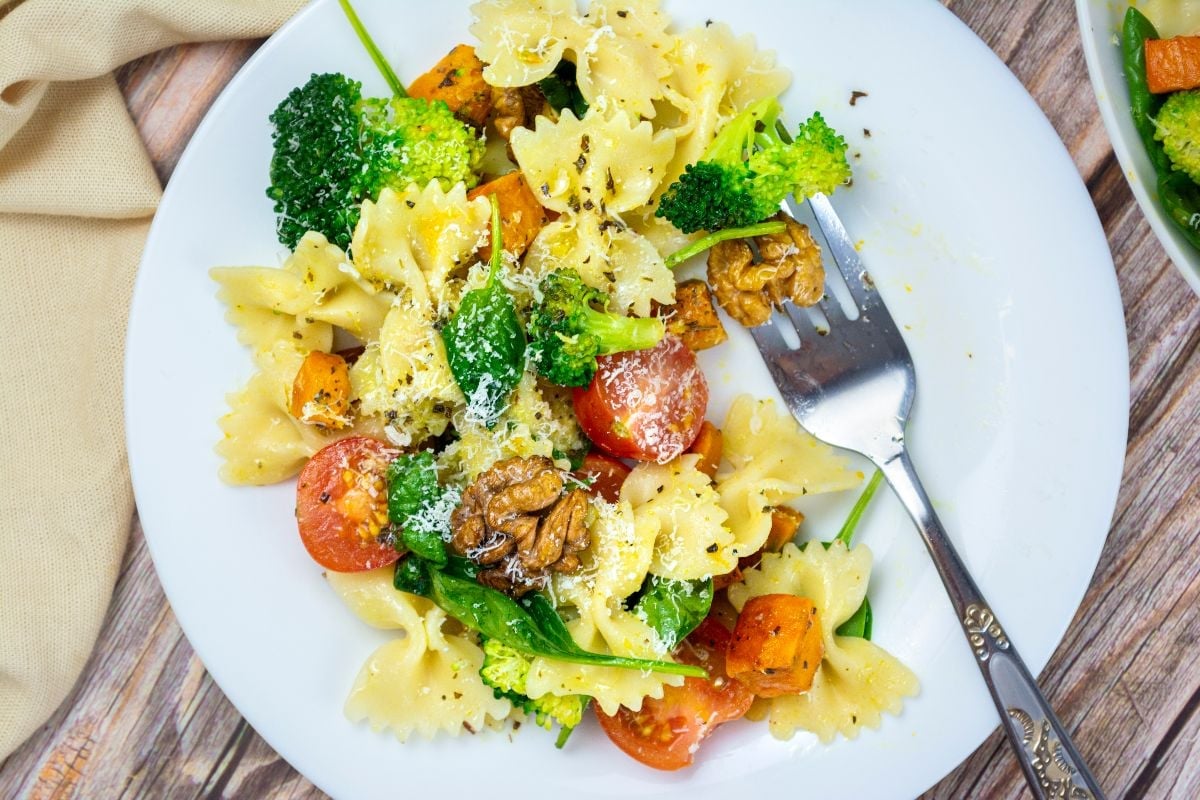 A plate of farfalle pasta with broccoli, cherry tomatoes, spinach, walnuts, grated cheese, and a fork on the side.