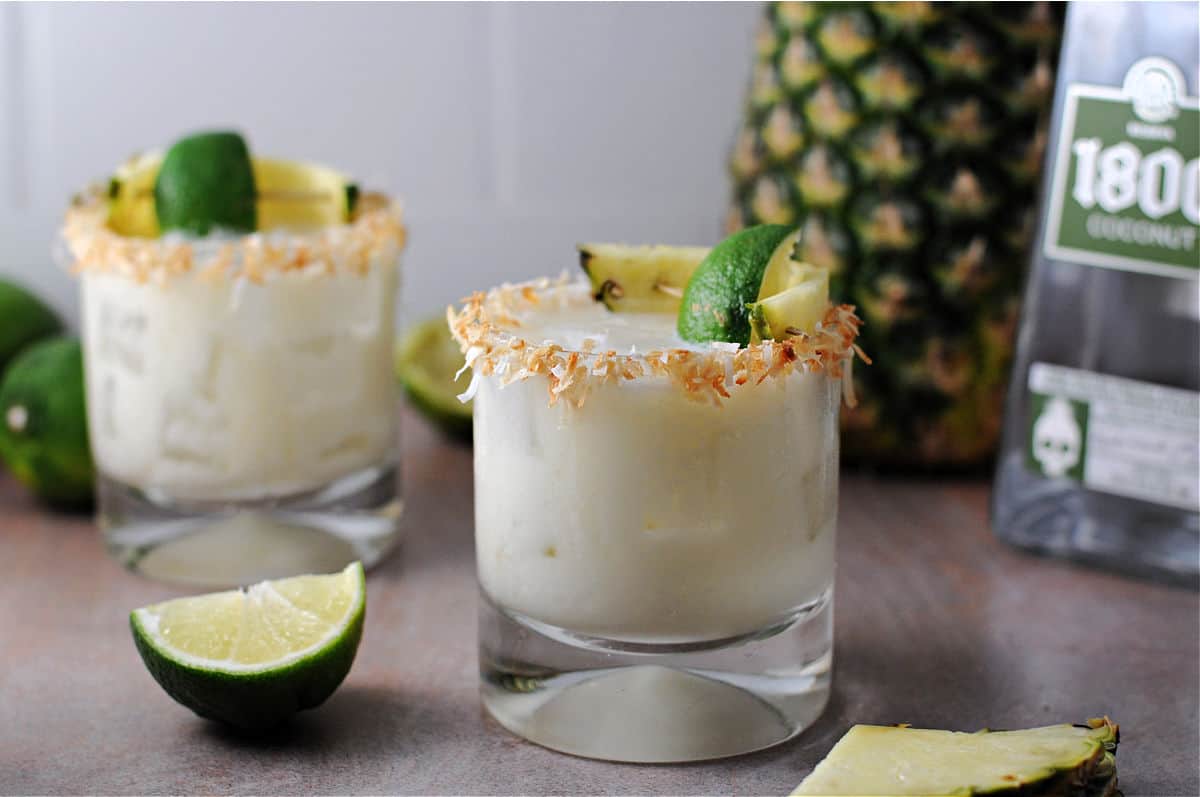 Two glasses of a creamy cocktail garnished with toasted coconut, lime wedges, and pineapple, with a bottle, limes, and a pineapple in the background.