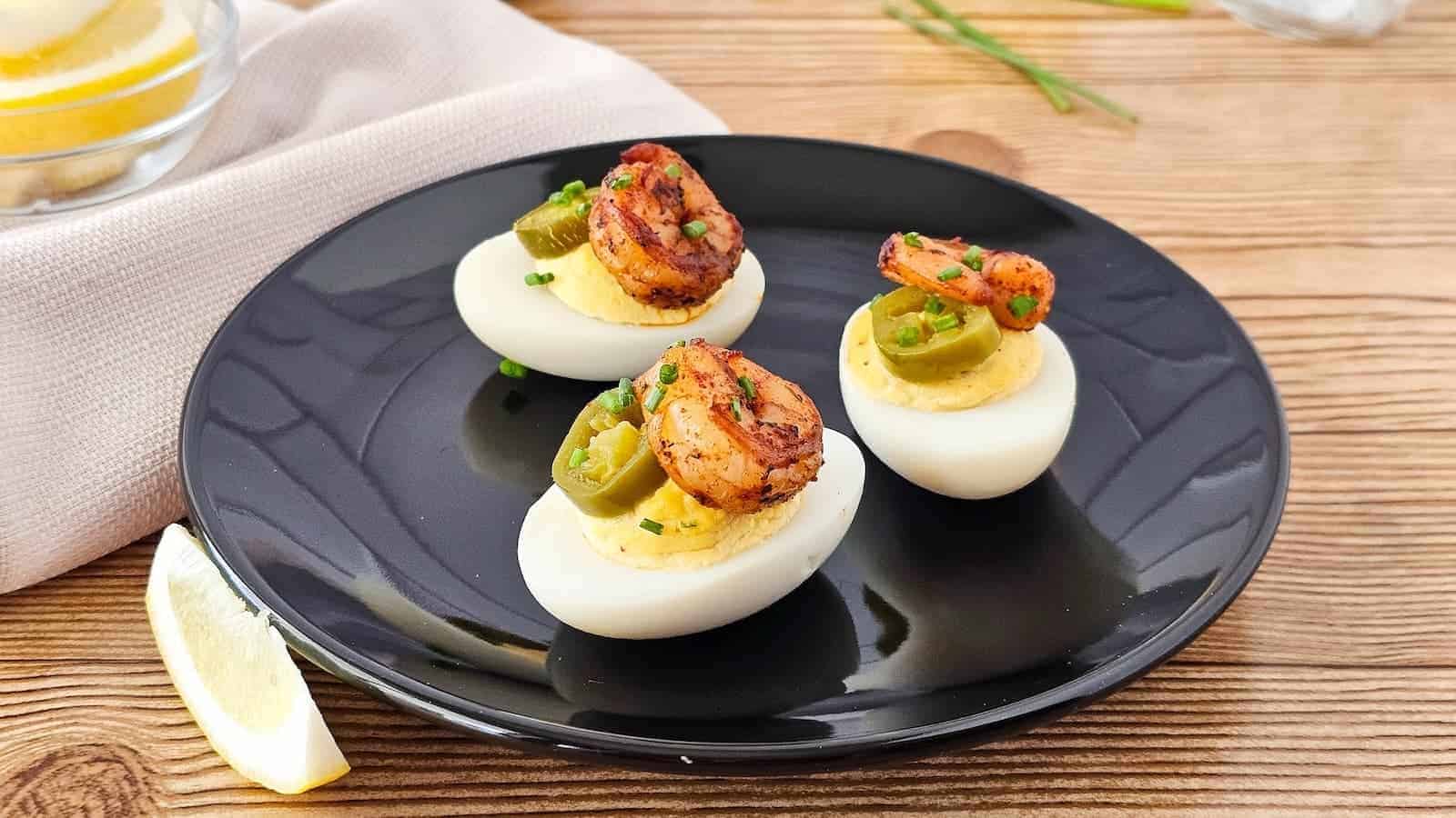 Three deviled eggs topped with grilled shrimp, jalapeno slices, and chopped chives are arranged on a black plate, with a lemon wedge beside the plate on a wooden surface.