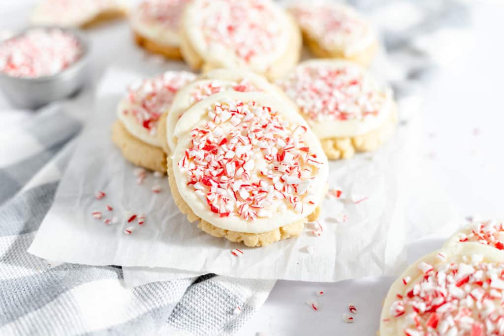 Sugar cookies topped with white frosting and crushed peppermint candies, arranged on parchment paper over a plaid cloth.