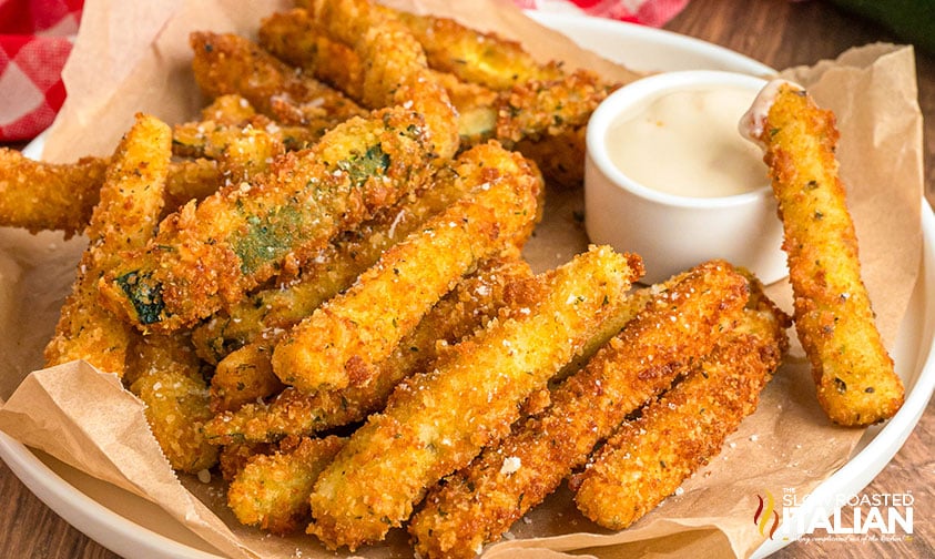 A plate of breaded and fried zucchini sticks is served with a small cup of dipping sauce on the side.