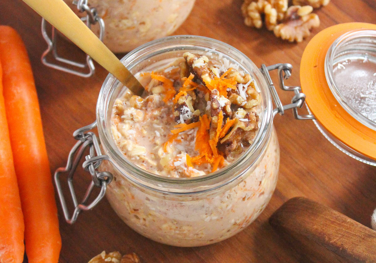 A glass jar filled with carrot cake overnight oats, topped with grated carrots, chopped walnuts, and shredded coconut, sits on a wooden surface next to carrots and walnut pieces.