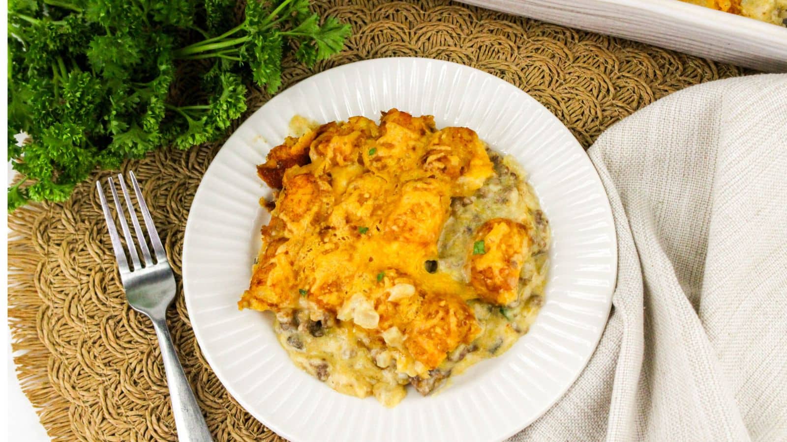 A serving of cheesy tater tot casserole with ground beef and creamy sauce on a white plate, placed on a woven placemat beside a fork, napkin, and fresh parsley.
