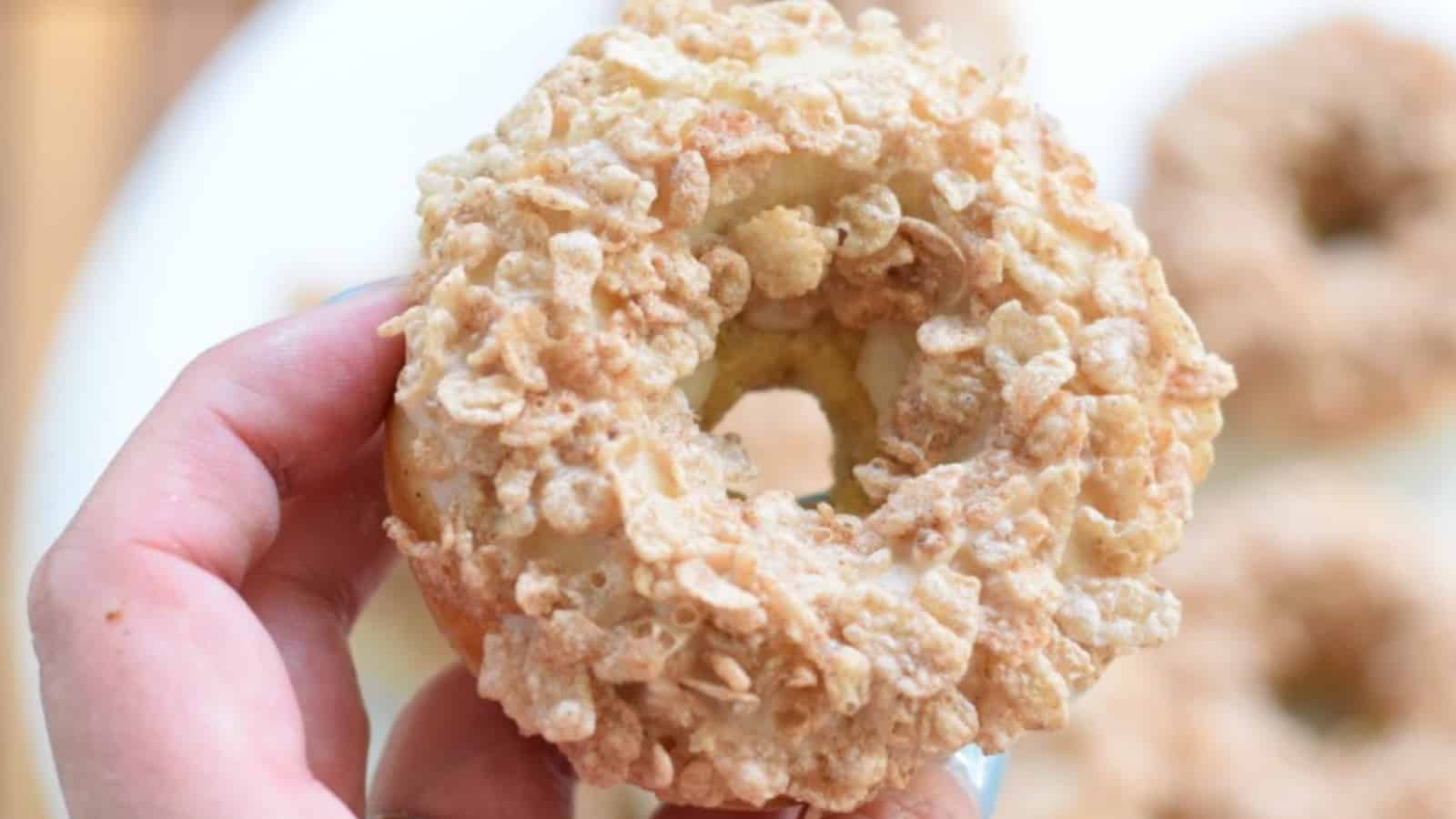 A hand holding a round doughnut covered in crispy cereal flakes, with a blurred plate of similar doughnuts in the background.