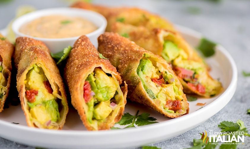 A plate of crispy avocado egg rolls cut in half, showing a filling of avocado, tomatoes, and herbs, with a dipping sauce in the background.