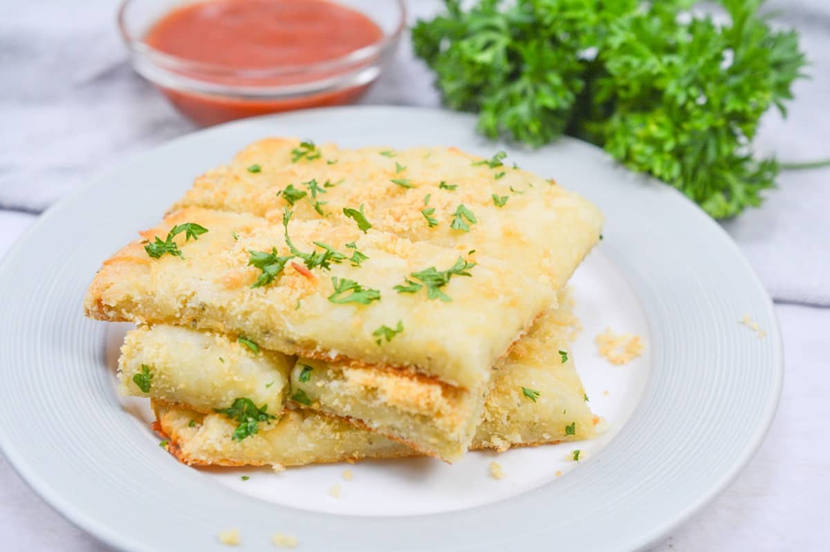 Three pieces of cheesy garlic bread garnished with parsley are stacked on a white plate, with a bowl of marinara sauce and fresh parsley in the background.