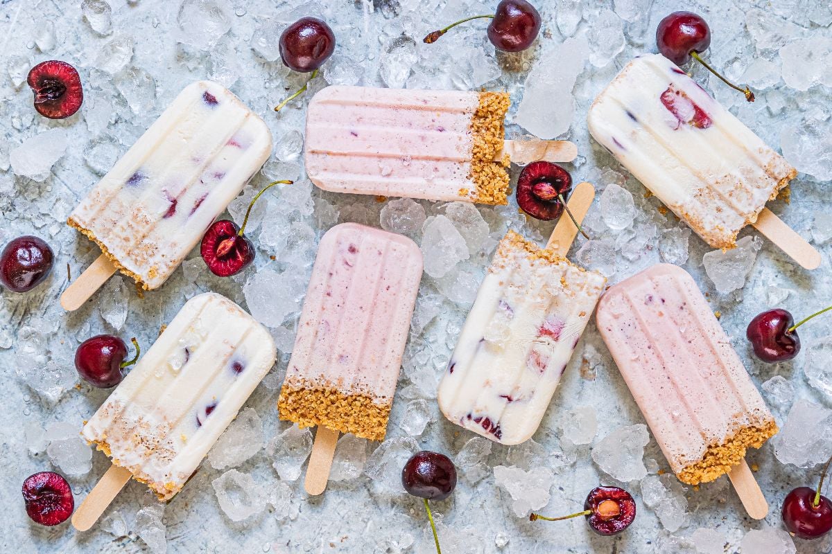 Eight creamy popsicles with cookie crumb coating are arranged on ice with scattered cherries.