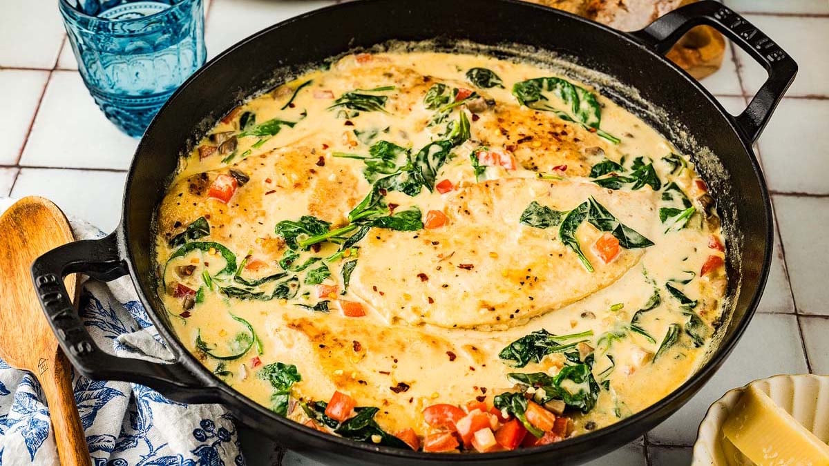 A skillet with creamy chicken breasts, spinach, and diced tomatoes in a rich sauce, displayed on a tiled surface with a wooden spoon nearby.