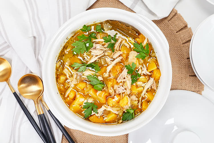 A white bowl filled with chicken soup containing shredded chicken, diced vegetables, and garnished with fresh parsley, placed on a burlap mat with spoons and plates nearby.