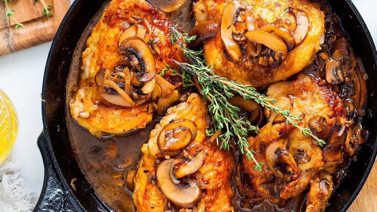 A cast iron skillet filled with cooked chicken thighs topped with sautรฉed mushrooms and garnished with fresh thyme.