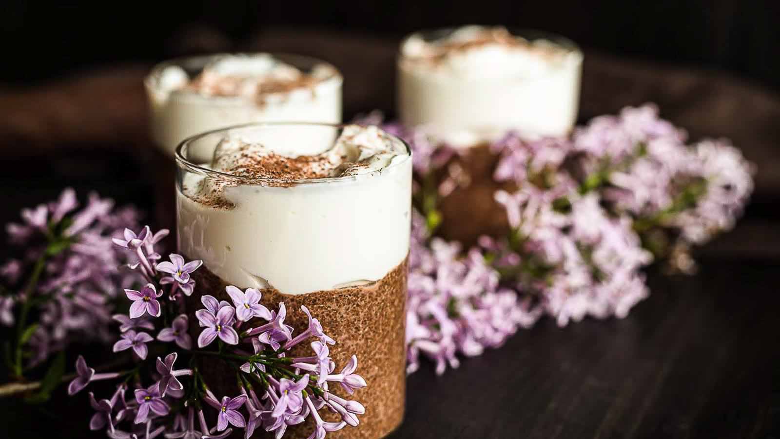 Three dessert glasses with layered chocolate and cream, garnished with cocoa powder and decorated with light purple lilac flowers on a dark surface.
