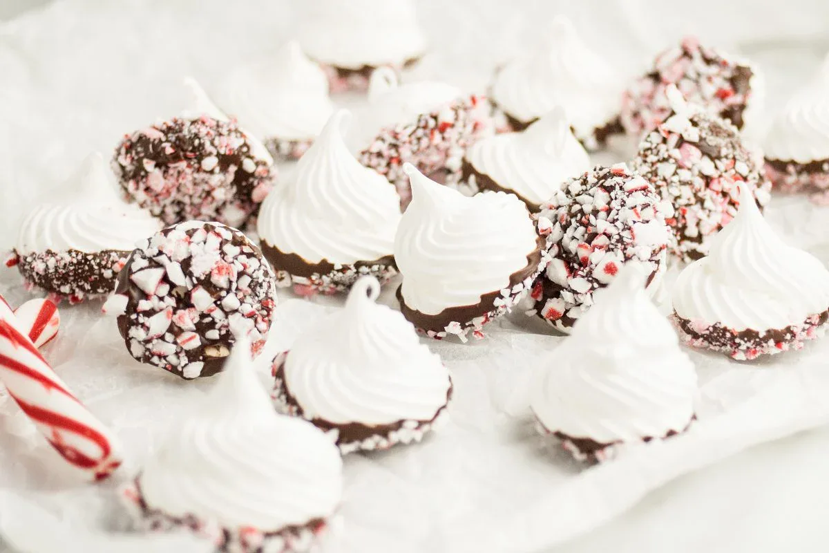Meringue cookies with peaks, some dipped in chocolate and coated with crushed peppermint candies, arranged on white parchment paper.