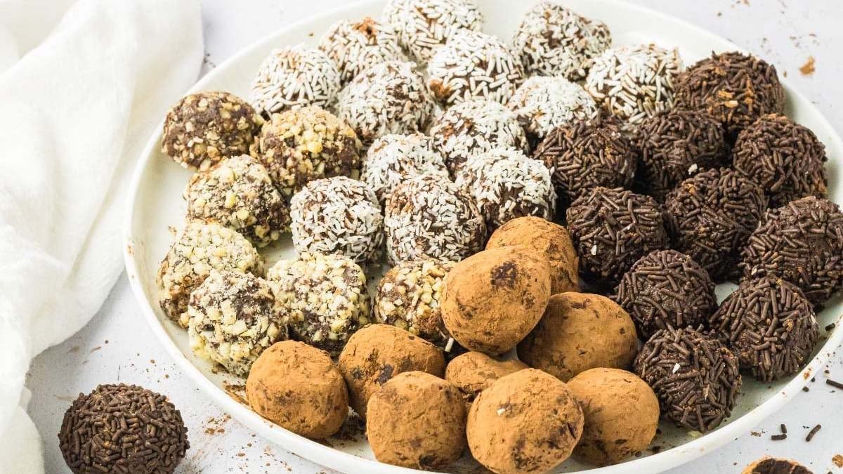 A white plate filled with assorted chocolate truffles coated in cocoa powder, chopped nuts, shredded coconut, and chocolate sprinkles.