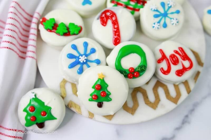 A plate of white chocolate-covered cookies decorated with colorful Christmas-themed icing designs, including trees, snowflakes, a wreath, and the word Joy.