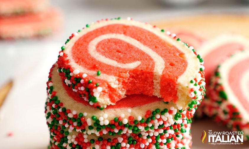 A stack of red and white swirl cookies coated with red, white, and green sprinkles; the top cookie has a bite taken out.