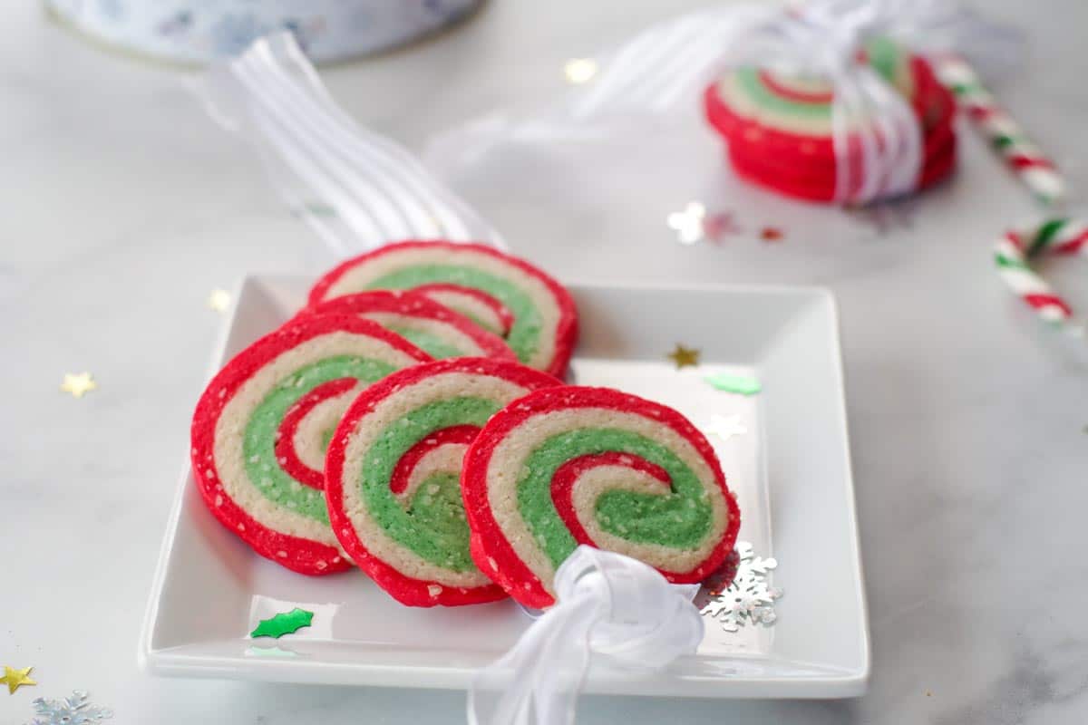 A square white plate with three round, red, white, and green swirl cookies. A fourth cookie is tied with a white ribbon in the background. Holiday confetti is scattered around.