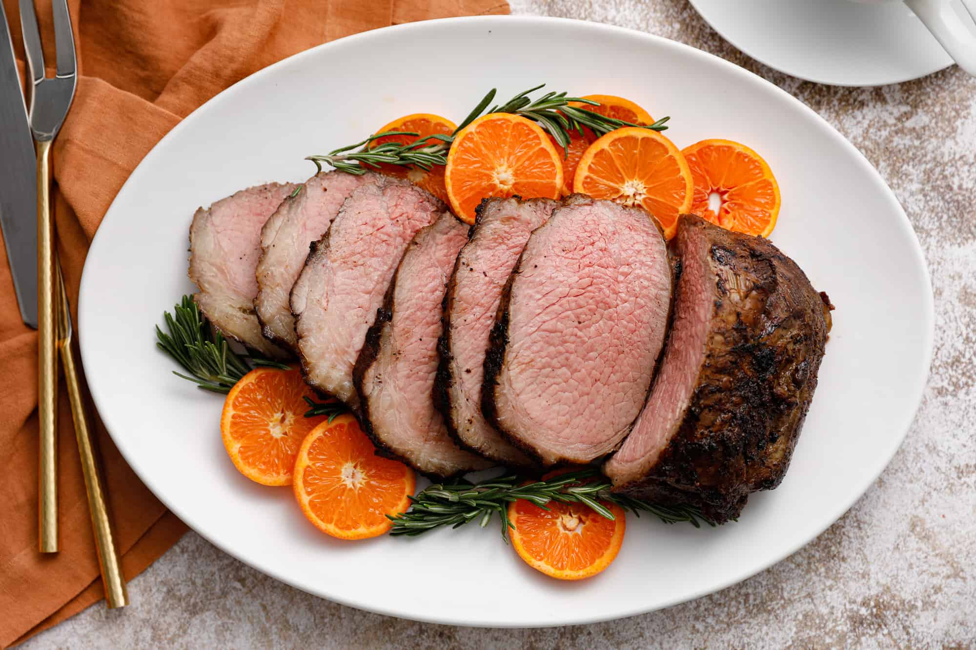 Sliced roast beef arranged on a white platter with fresh rosemary sprigs and halved oranges for garnish.