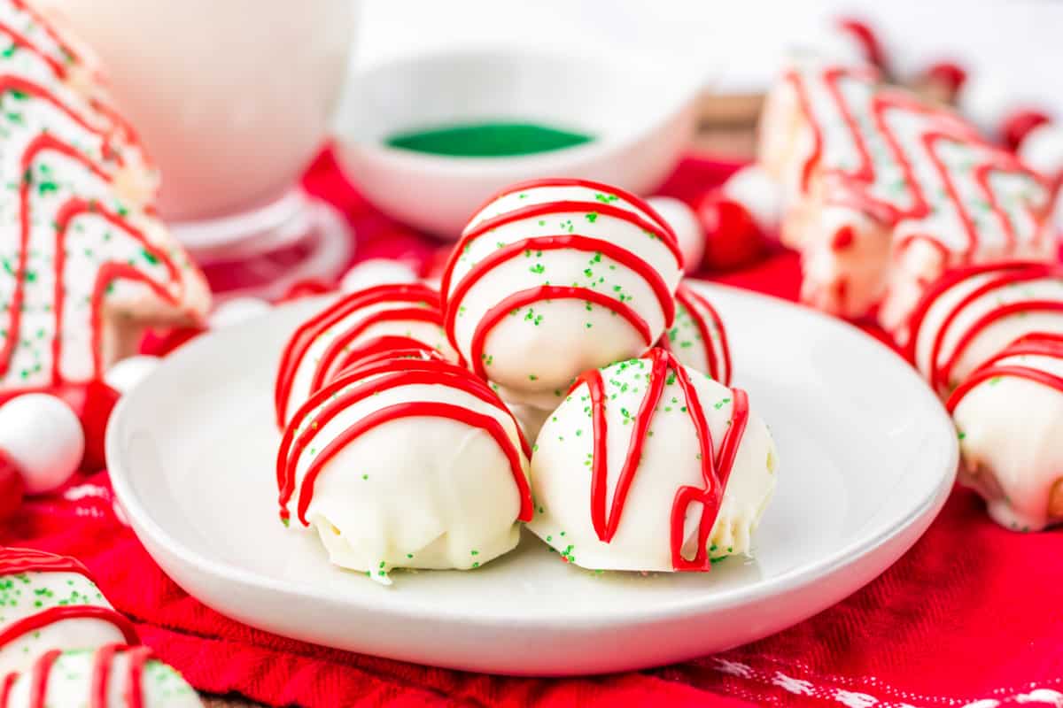 A white plate with round, white chocolate-covered treats drizzled with red icing and sprinkled with green sugar, set on a red cloth.
