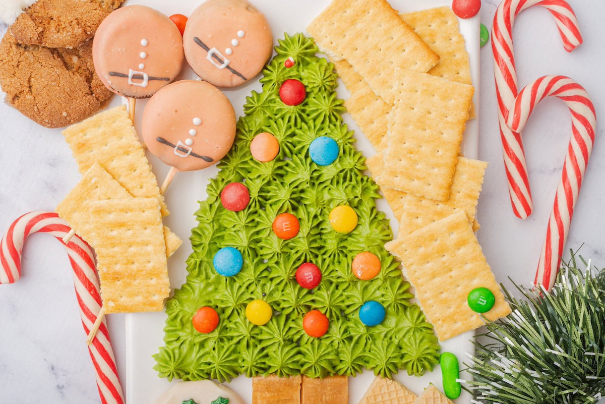 A platter with green frosting shaped like a Christmas tree, decorated with colorful candies, surrounded by crackers, cookies, and candy canes.