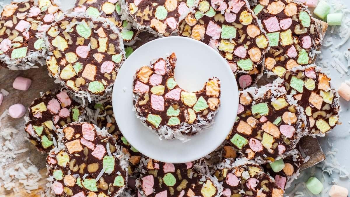 Sliced dessert made of multicolored mini marshmallows and chocolate, coated with shredded coconut, arranged on a surface with one piece on a white plate showing a bite taken out.