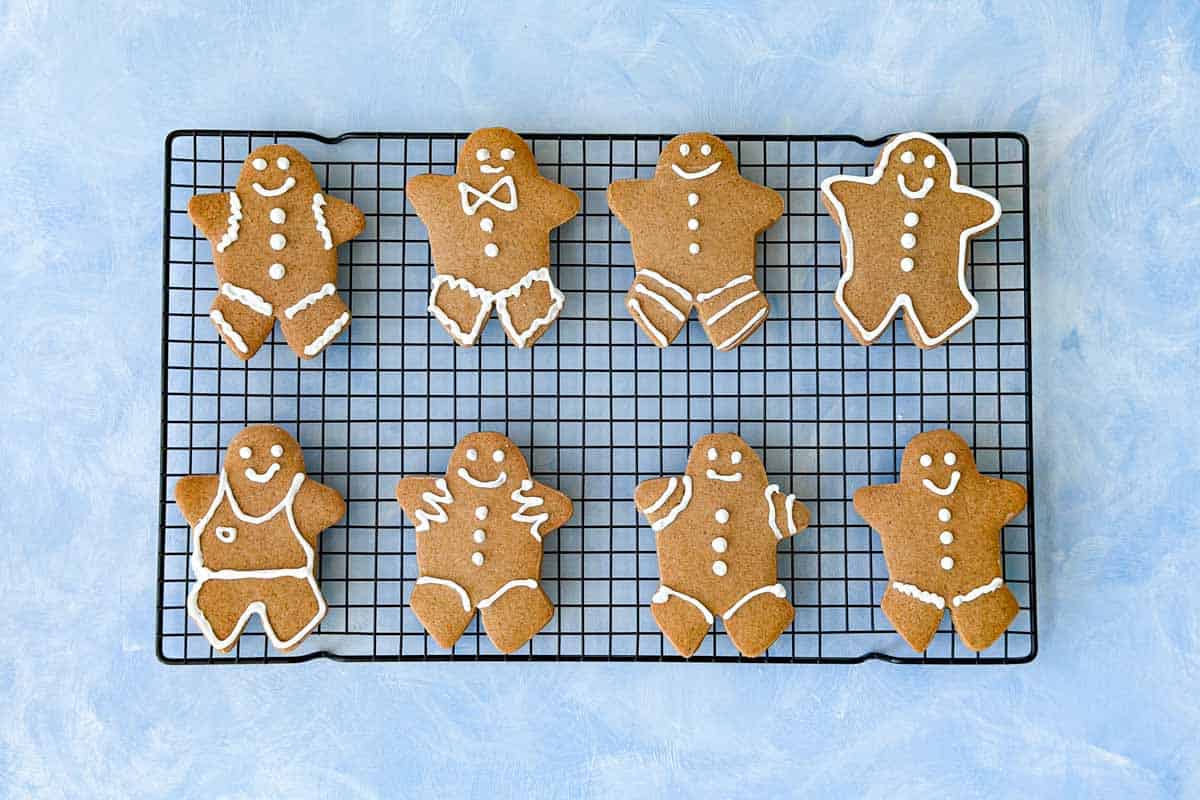 Eight gingerbread cookies decorated with white icing are arranged on a black cooling rack against a light blue background.