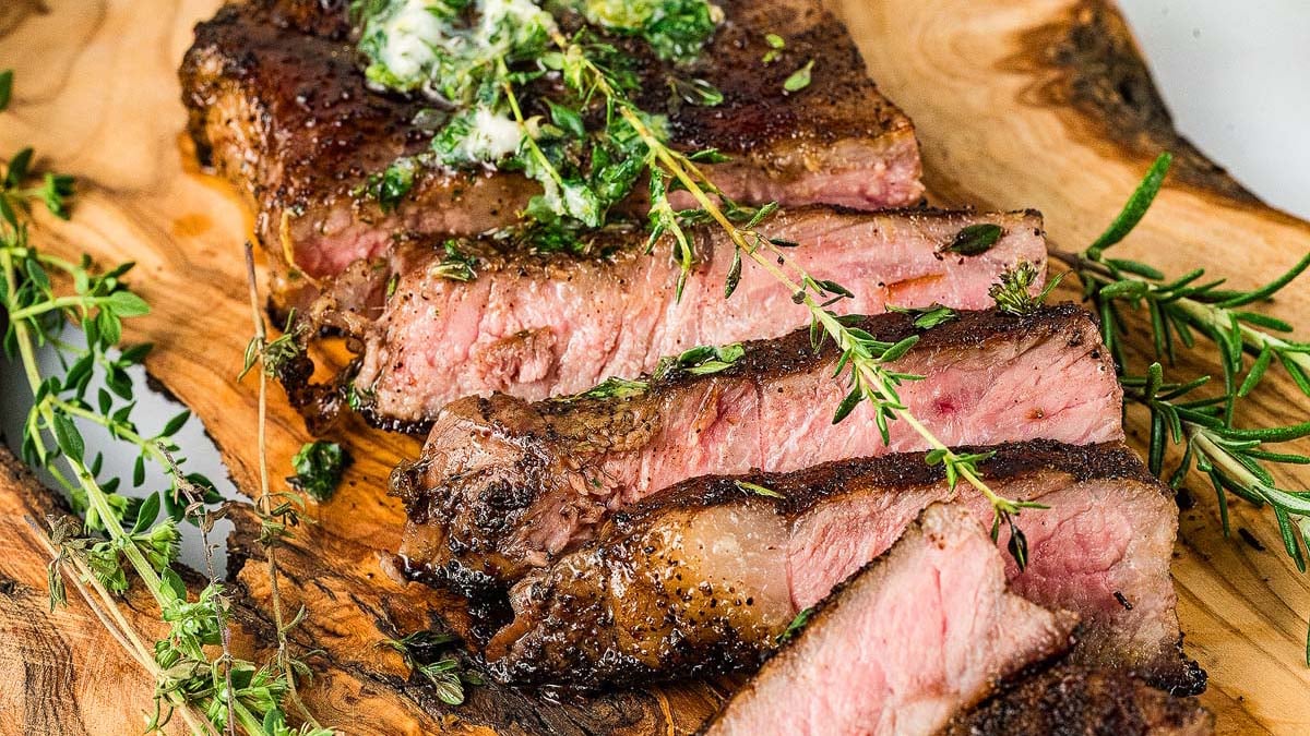 Sliced medium-rare steak garnished with herbs and melted herb butter on a wooden cutting board.