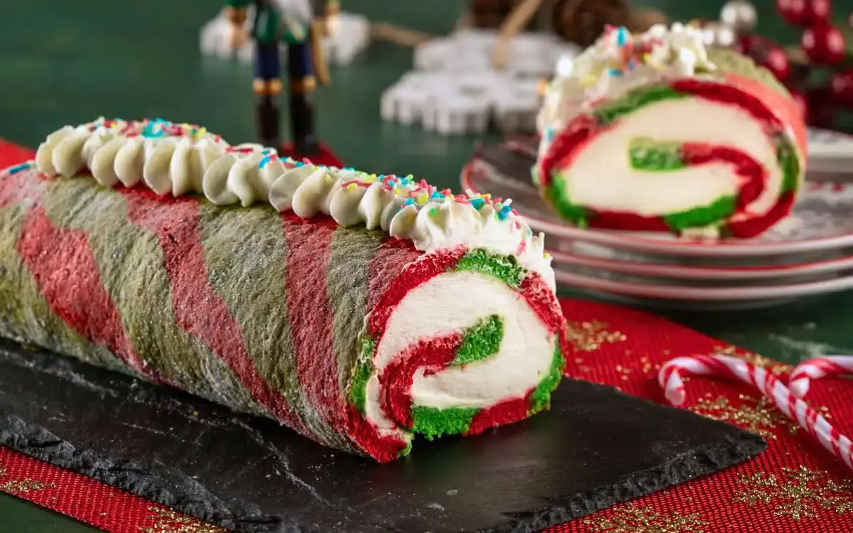 A festive red, green, and white rolled cake with swirled colors, topped with whipped cream and rainbow sprinkles, sits on a slate board with a slice on a plate in the background.